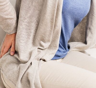 Person in gray cardigan and blue tank top holding lower back, suggesting back pain or discomfort